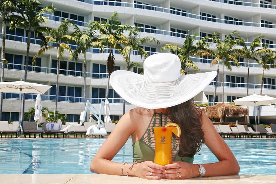 Woman in white hat at resort pool holding tropical drink with high-rise building backdrop