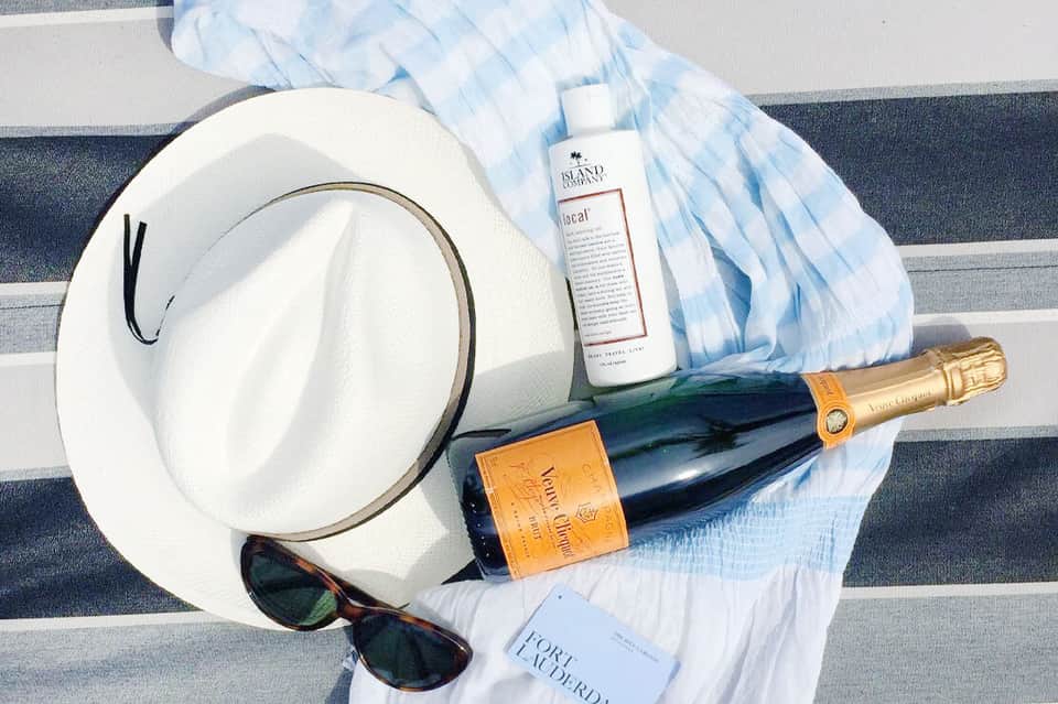 Poolside luxury items: white hat, champagne bottle, sunscreen, sunglasses on striped towel