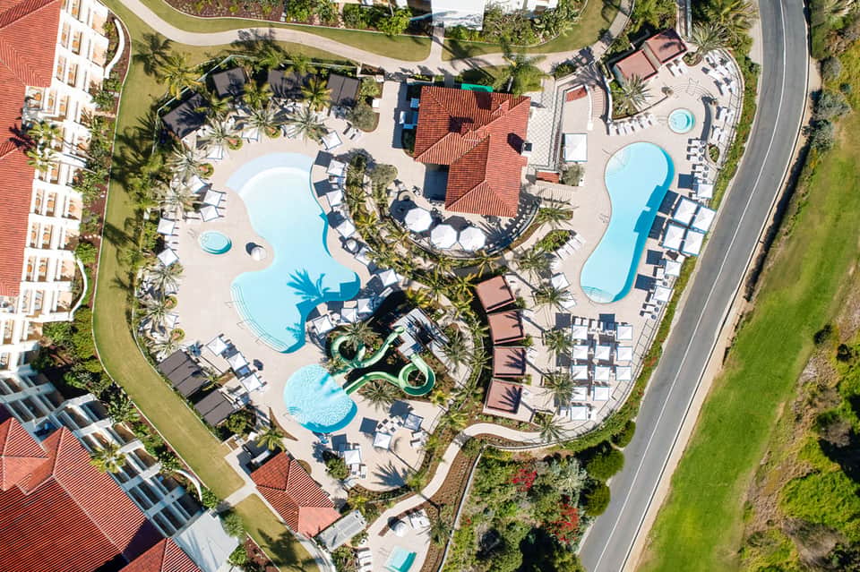Aerial view of resort with multiple curved pools, loungers, and tropical landscaping surrounded by buildings