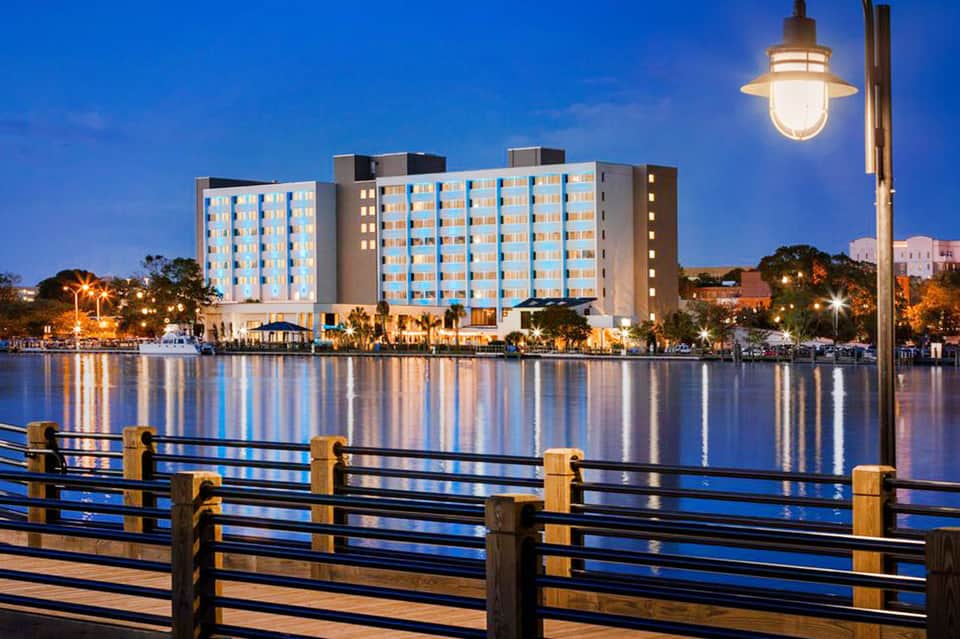 Illuminated hotel building reflected in calm water at dusk with wooden railing in foreground