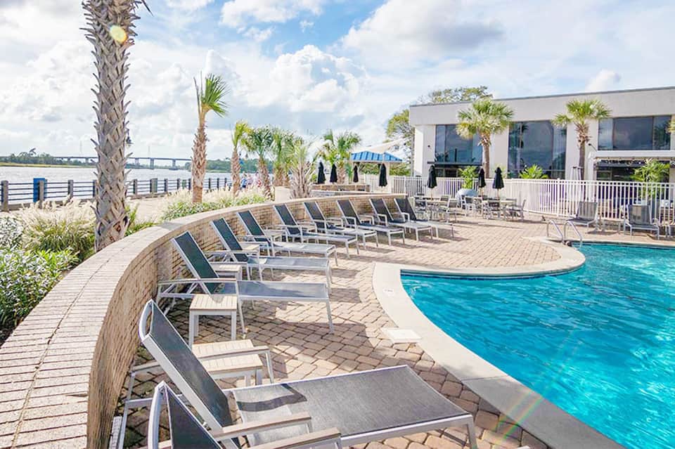 Curved poolside lounge chairs with palm trees and waterfront building in background