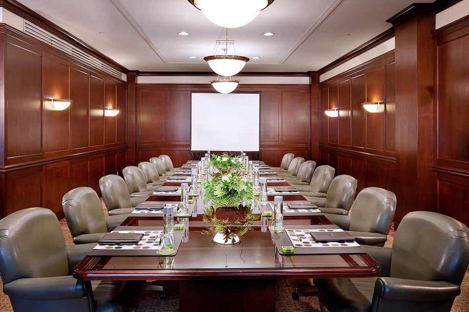 Executive boardroom with dark wood paneling, long conference table, leather chairs, and projection screen