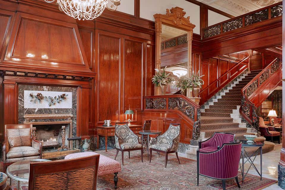 Elegant classical lobby with rich wood paneling, fireplace, ornate staircase, crystal chandelier, and period furniture
