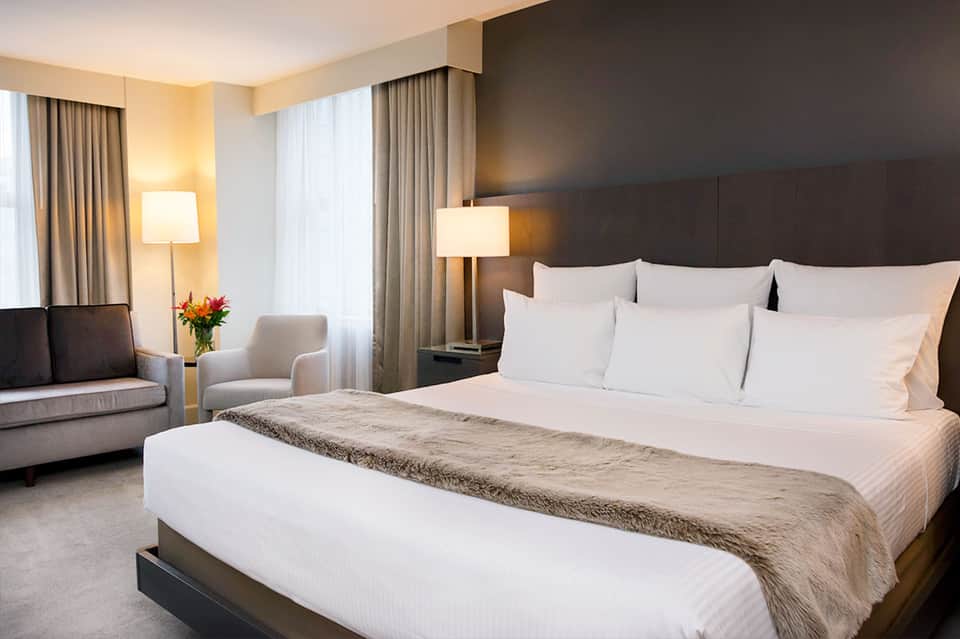Luxury hotel bedroom with king bed, dark headboard, sitting area, and warm lighting