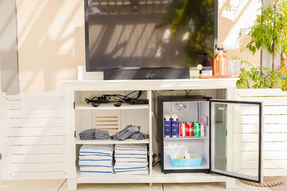 White outdoor entertainment cabinet with open door revealing mini refrigerator stocked with beverages and folded towels on shelves