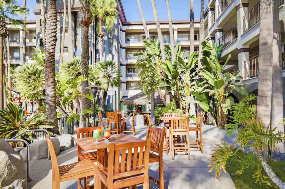 Tropical courtyard with palm trees, wooden dining tables, and multi-story hotel buildings surrounding the space