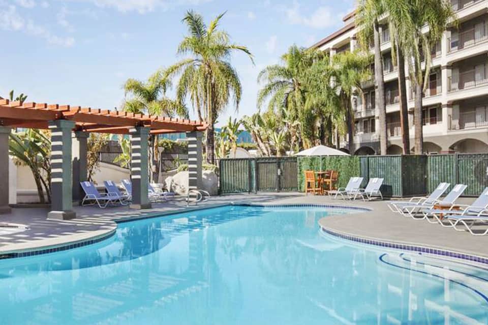 Resort pool with turquoise water, lounge chairs, pergola structures, and multi-story hotel building