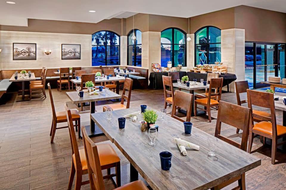 Modern restaurant dining room with wooden tables, orange chairs, and large arched windows overlooking the ocean