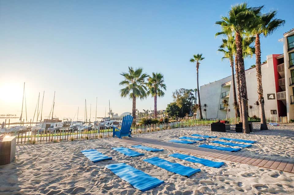 Beach yoga class with blue mats lined on sand near sailboats and palm trees