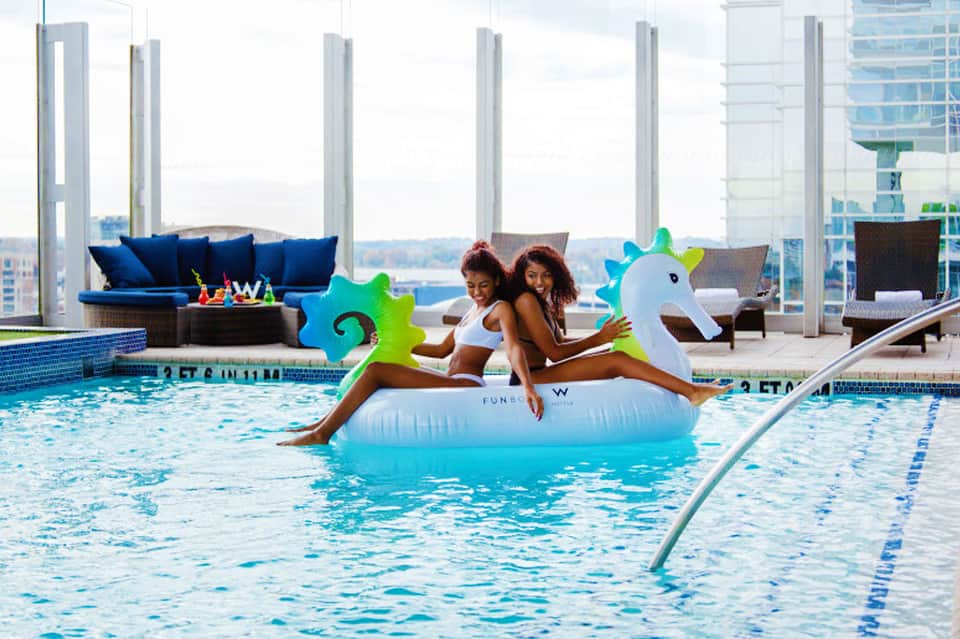 Indoor pool with two women on a unicorn float float against a city skyline backdrop