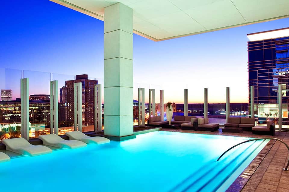 Rooftop infinity pool at dusk with glowing turquoise water, city skyline, lounge seating, and support columns