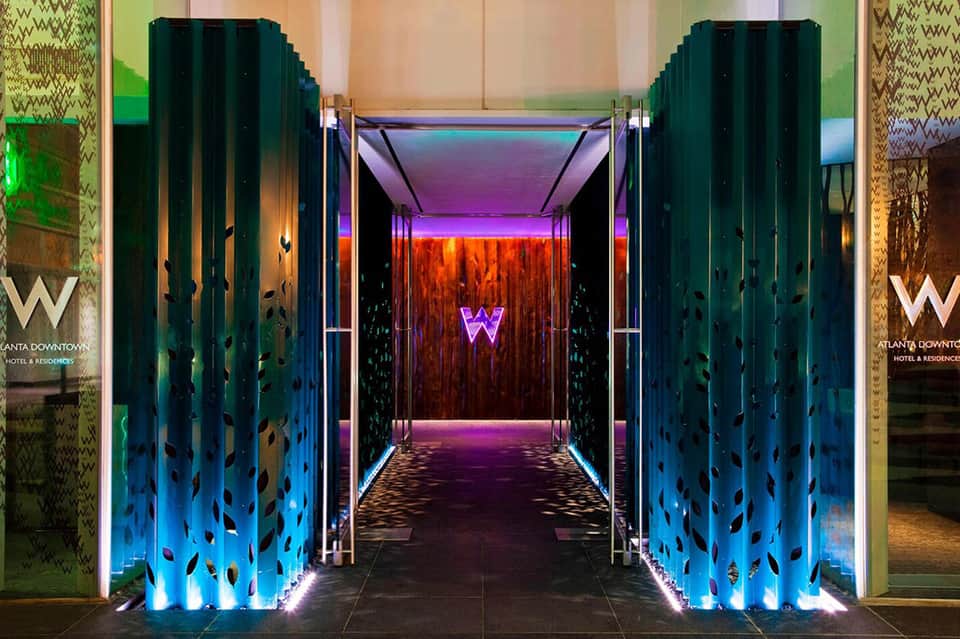 Illuminated hotel corridor entrance with dark teal perforated doors, blue and orange neon lighting, and patterned wallpaper
