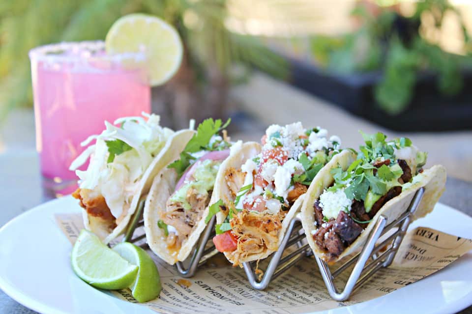 Four gourmet tacos with lime and pink drink on white plate with garnish