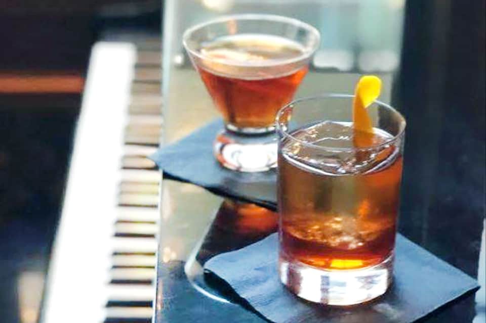 Two cocktails on a piano keyboard - amber drink with lemon twist and dark cocktail with foam top