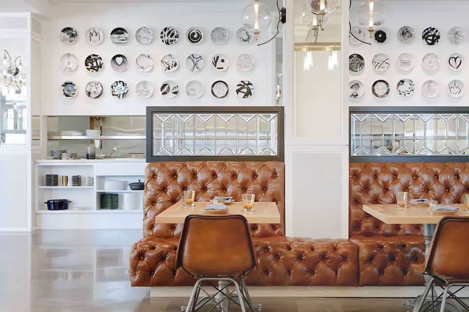 Modern dining area with tufted cognac leather banquette, decorative plate wall display, and geometric glass-fronted cabinetry.
