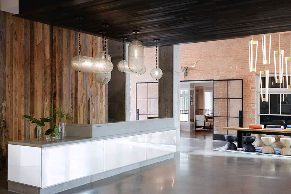 Modern hotel lobby with white reception desk, wooden accent wall, hanging pendant lights, and brick architecture