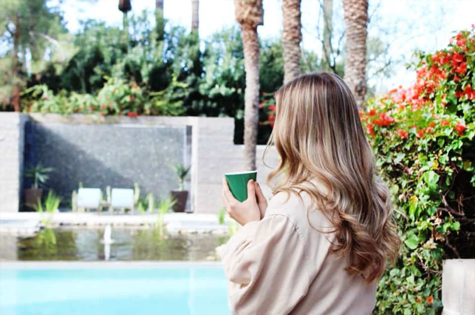Woman in beige jacket holding green cup overlooking resort pool surrounded by palm trees and landscaping