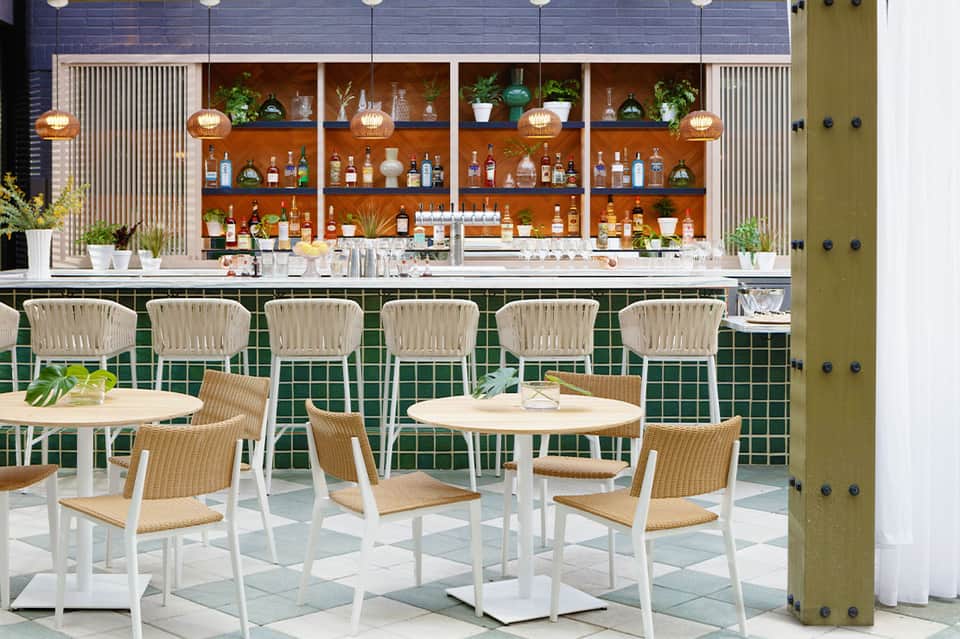 Modern bar lounge with green tile walls, wooden shelving displaying bottles, and woven bar stools