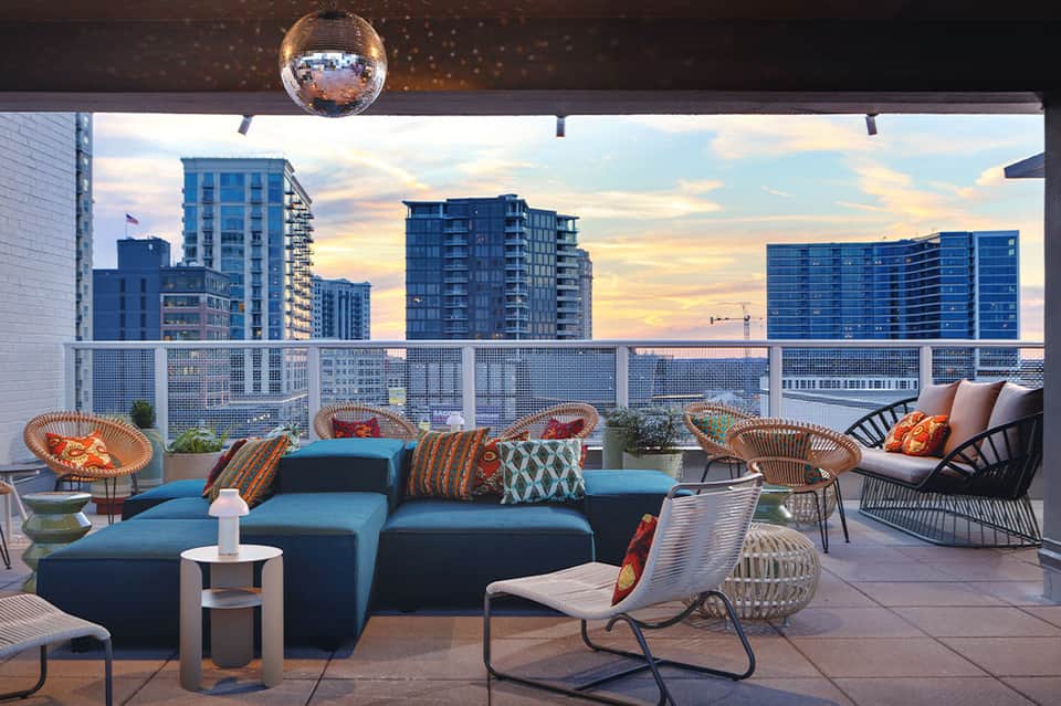 Modern rooftop lounge with blue seating, wicker chairs, and cityscape views at sunset with disco ball overhead