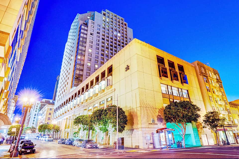 Elegant historic building with cream facade and large windows illuminated at dusk, surrounded by modern high-rise hotels