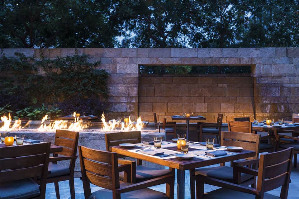 Outdoor dining area with wooden tables and chairs beside fire pits and stone wall at dusk