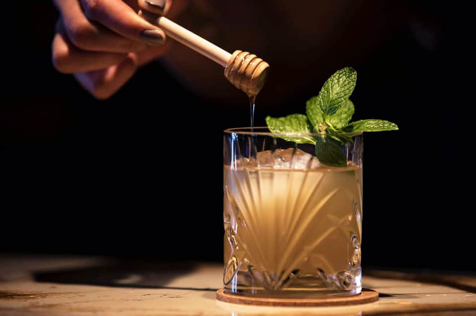 Craft cocktail being prepared with honey drizzle, fresh mint garnish, and golden spirits in a rocks glass