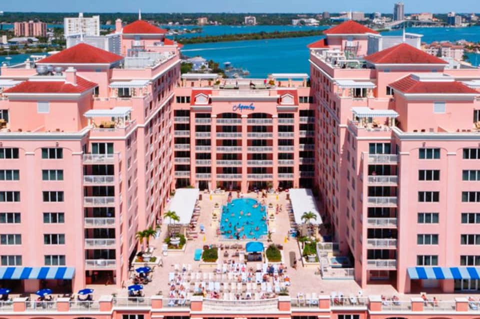 Aerial view of pink resort with central courtyard pool, red-roofed buildings, and turquoise ocean backdrop