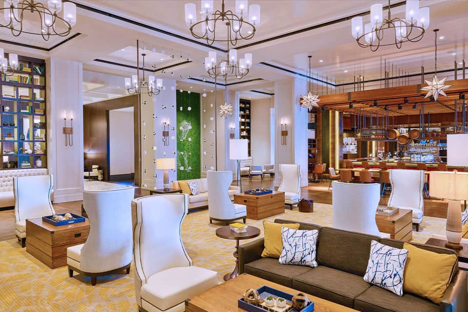 Elegant hotel lounge with white upholstered chairs, wooden tables, crystal chandeliers, and a modern bar area in background