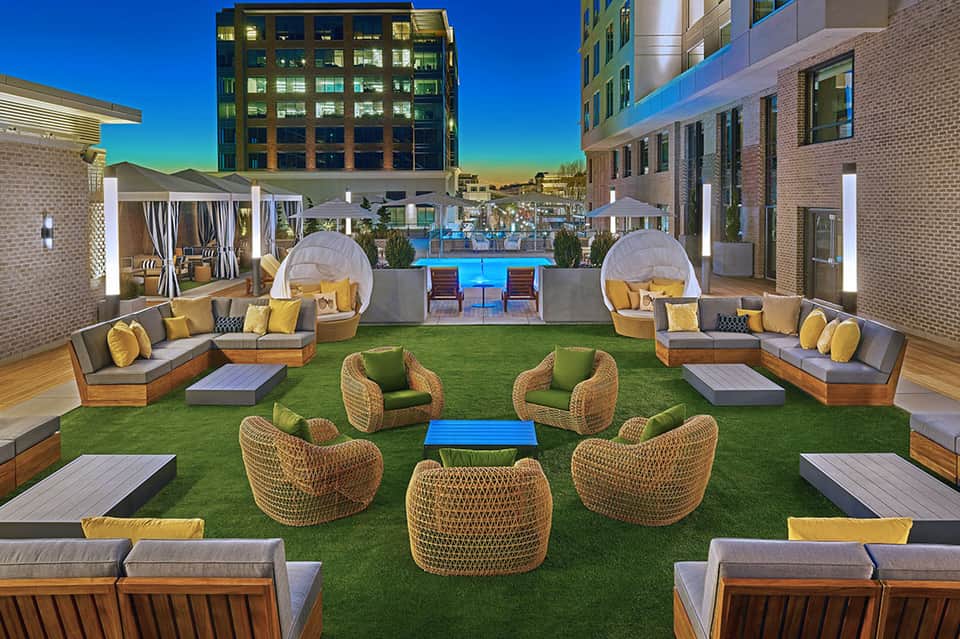 Modern rooftop terrace with glowing cabanas, woven chairs, artificial turf, and illuminated pool at dusk