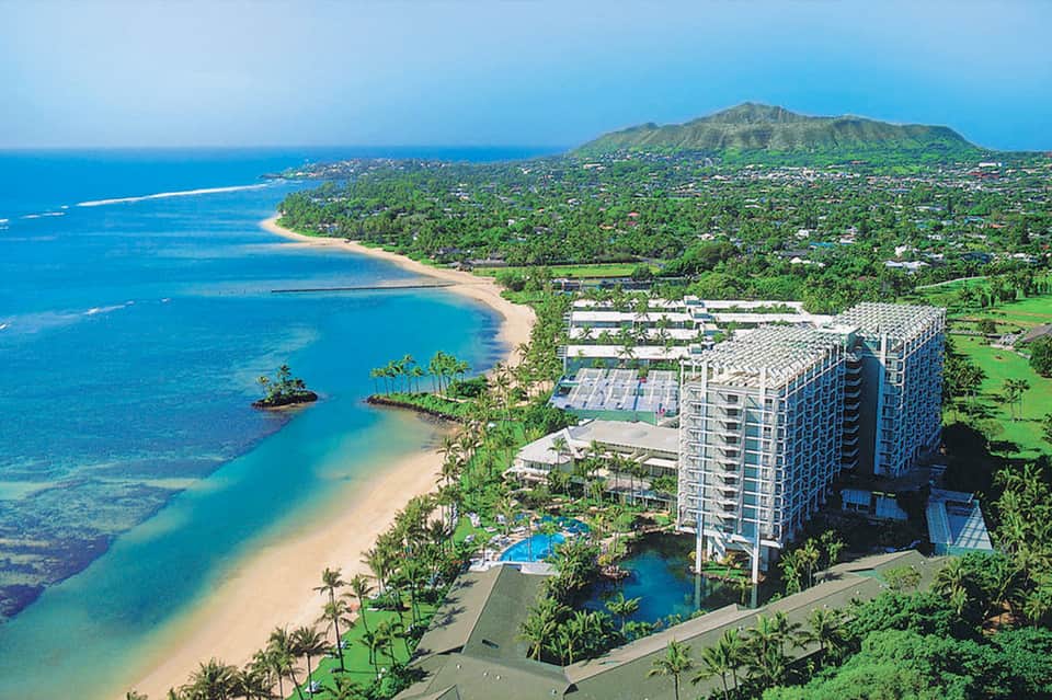 Aerial view of beachfront resort with high-rise hotel, pools, and lush tropical landscaping along blue ocean