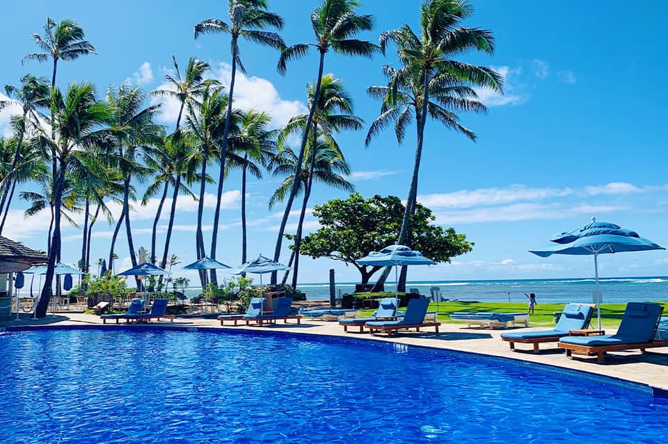 Resort pool with palm trees, blue umbrellas, lounge chairs, and ocean view in tropical setting