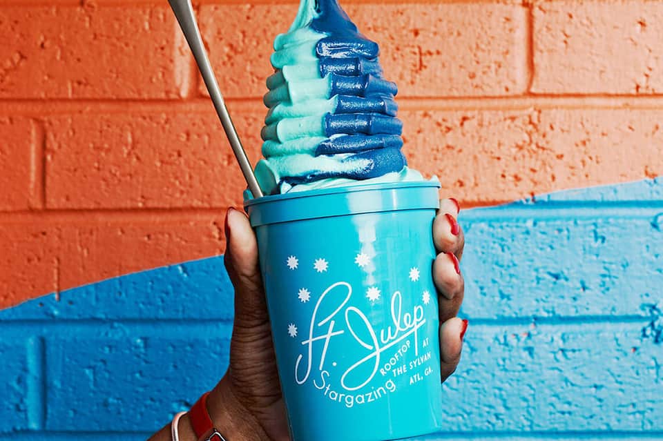 Hand holding turquoise cup with blue and white swirled frozen treat against painted brick wall