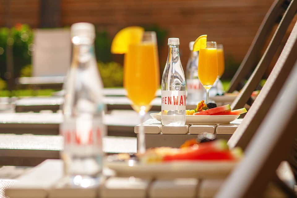 Outdoor rooftop dining setup with orange juice cocktails, fresh vegetables, and water bottles on a sunny terrace.