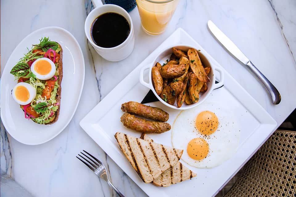 Gourmet breakfast spread with avocado toast, eggs, sausages, potatoes, coffee, and juice