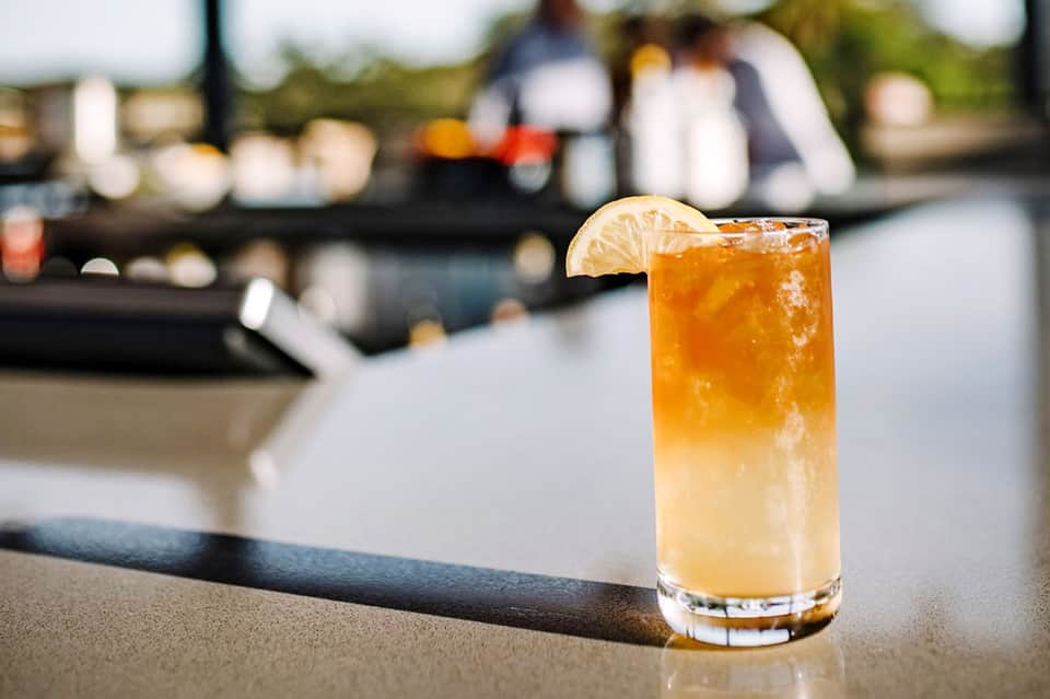 Amber cocktail with lime wedge garnish on outdoor bar counter with blurred waterfront setting