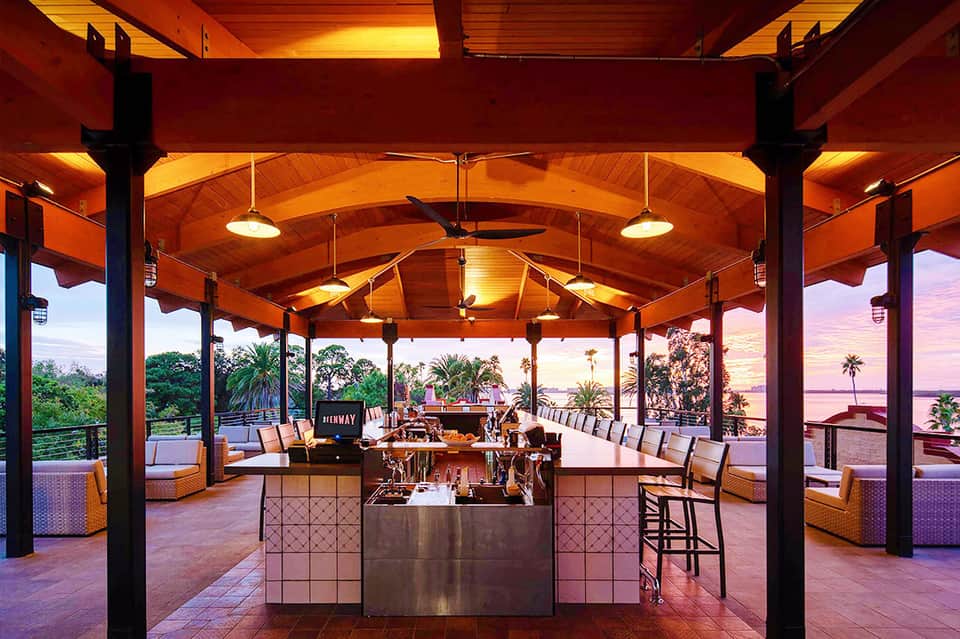 Elegant outdoor lounge pavilion with wooden beams, orange lighting, bar counter, and sunset ocean views