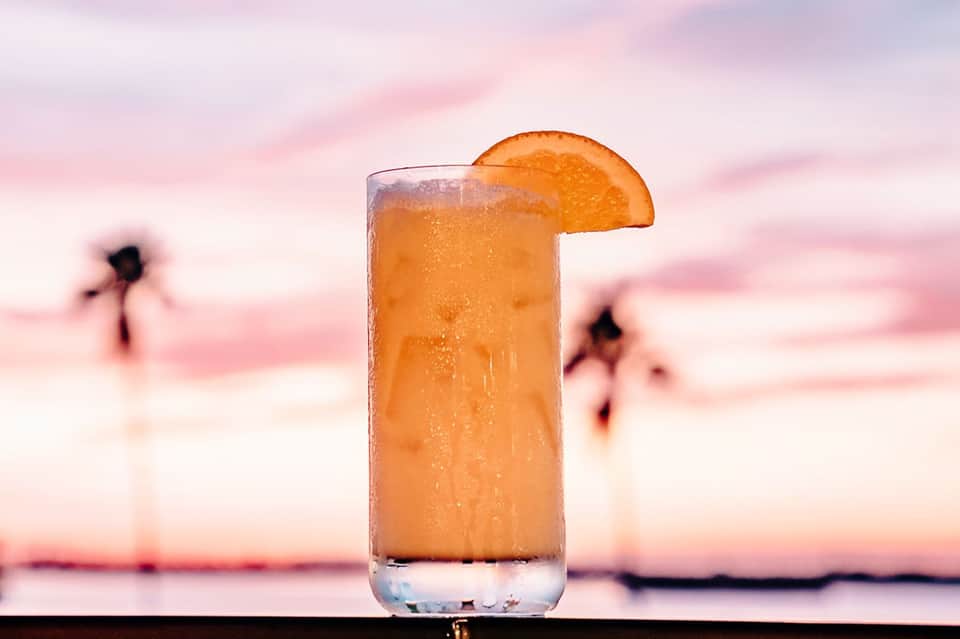 Orange cocktail with orange slice garnish at sunset beach with swimmers in background