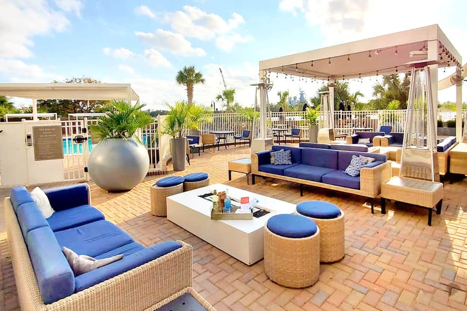 Modern rooftop lounge with blue seating, pergolas, fire pit, and pool view overlooking palm trees