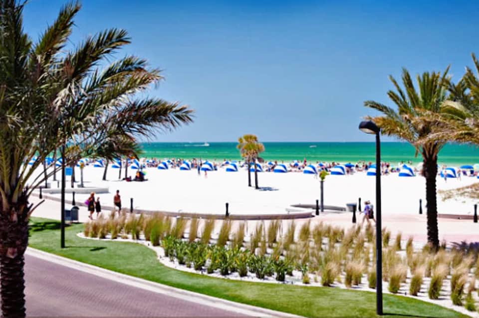 Beachfront resort with white sand, turquoise ocean, palm trees, and manicured coastal landscaping with beach umbrellas