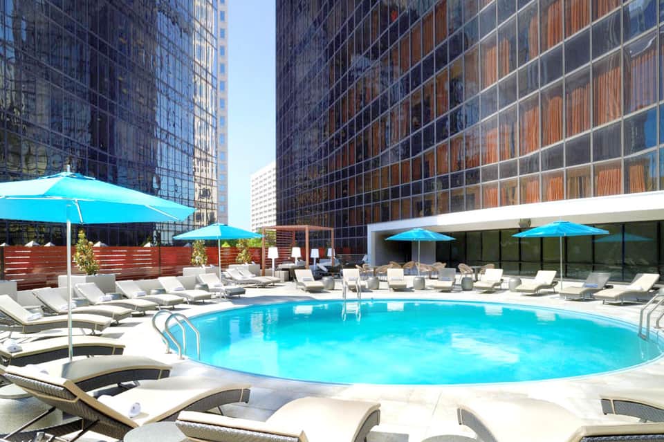 Urban rooftop pool with bright blue water, lounge chairs, blue umbrellas, and tall glass buildings