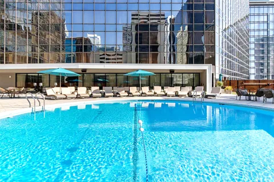 Olympic-sized swimming pool with turquoise water, lounge chairs, umbrellas, and city skyline backdrop