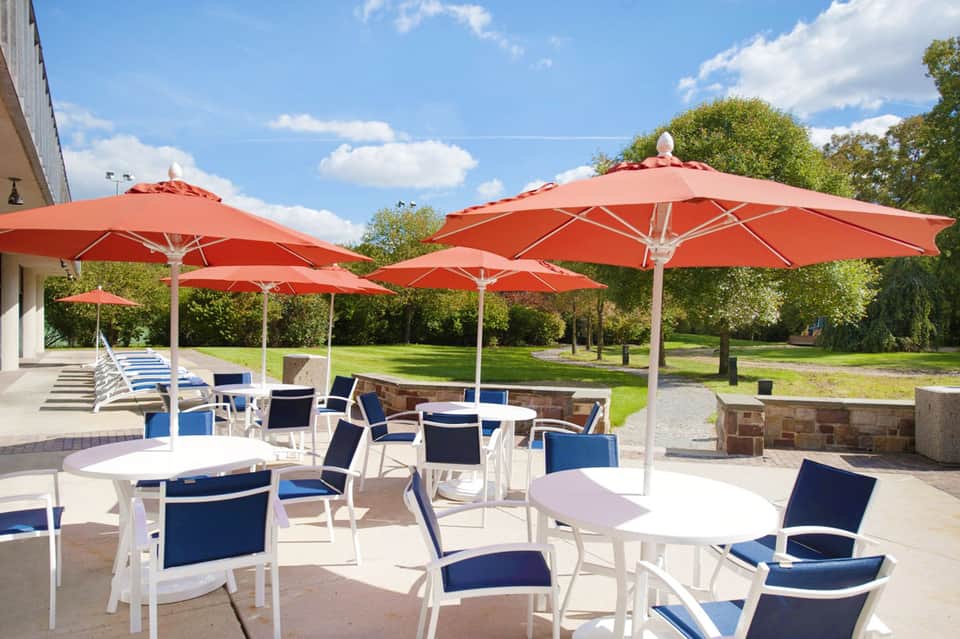 Outdoor patio with white tables, chairs, and red umbrellas overlooking green lawn and trees