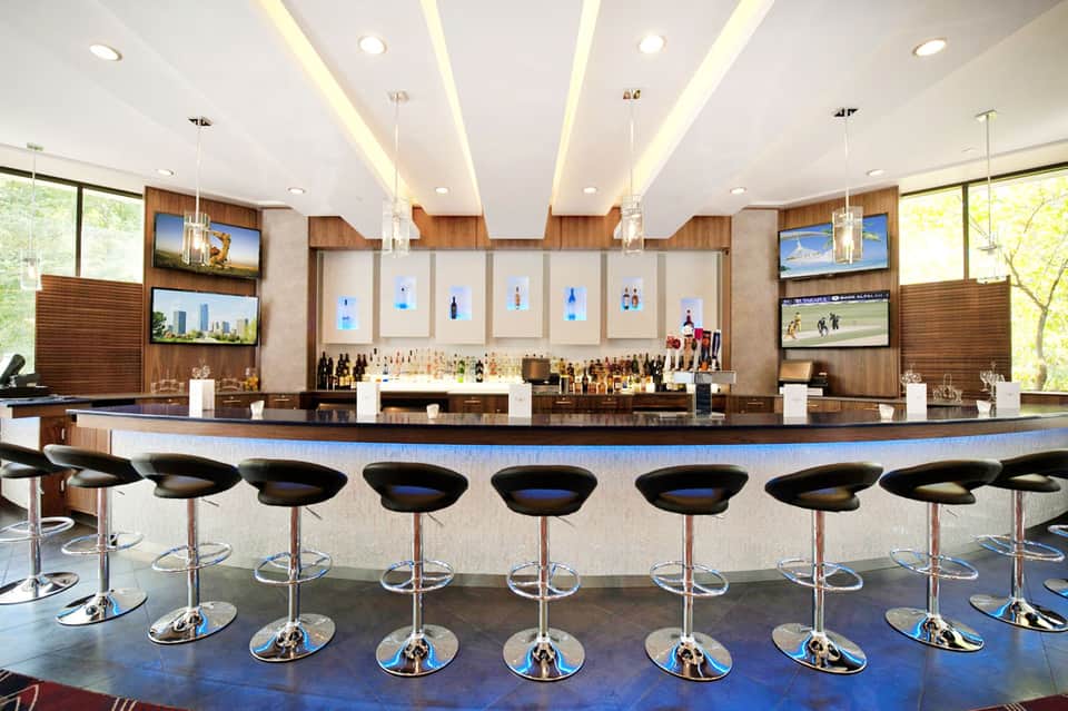 Modern hotel bar with curved counter, black stools, and illuminated ceiling with wood accents