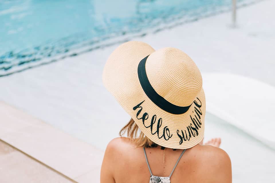 Woman wearing straw hat with 'hello sunshine' text by pool overlooking water