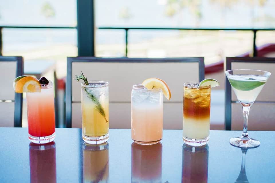 Five colorful cocktails on a bar counter with ocean view and beach lounge chairs visible