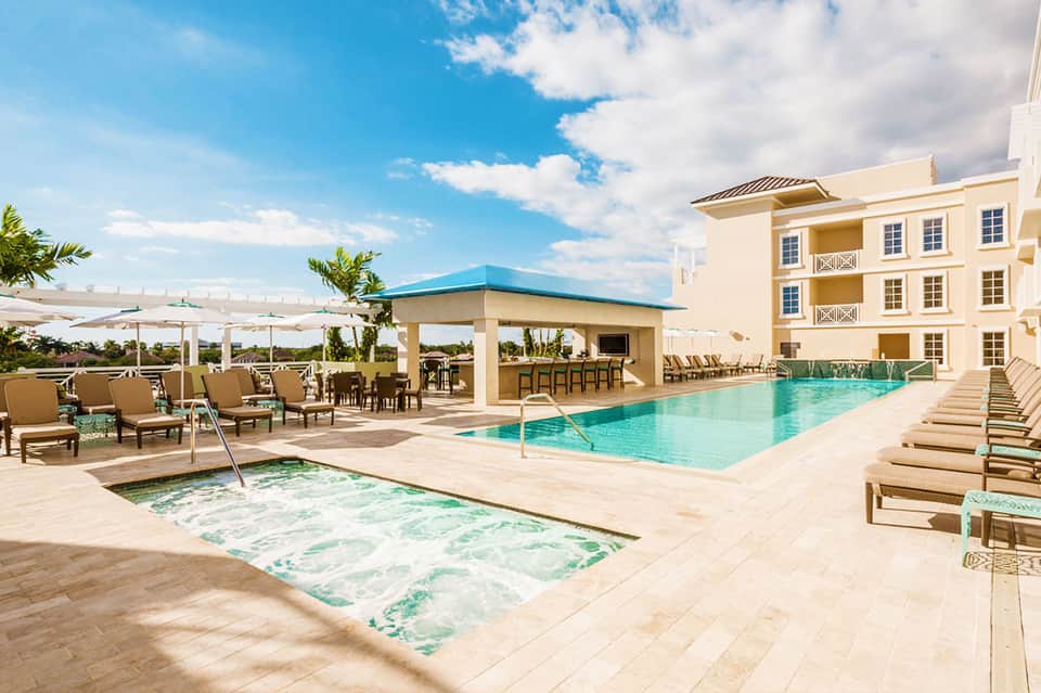 Resort pool area with spa tub, cabana, tan loungers, and multi-story yellow building under blue sky