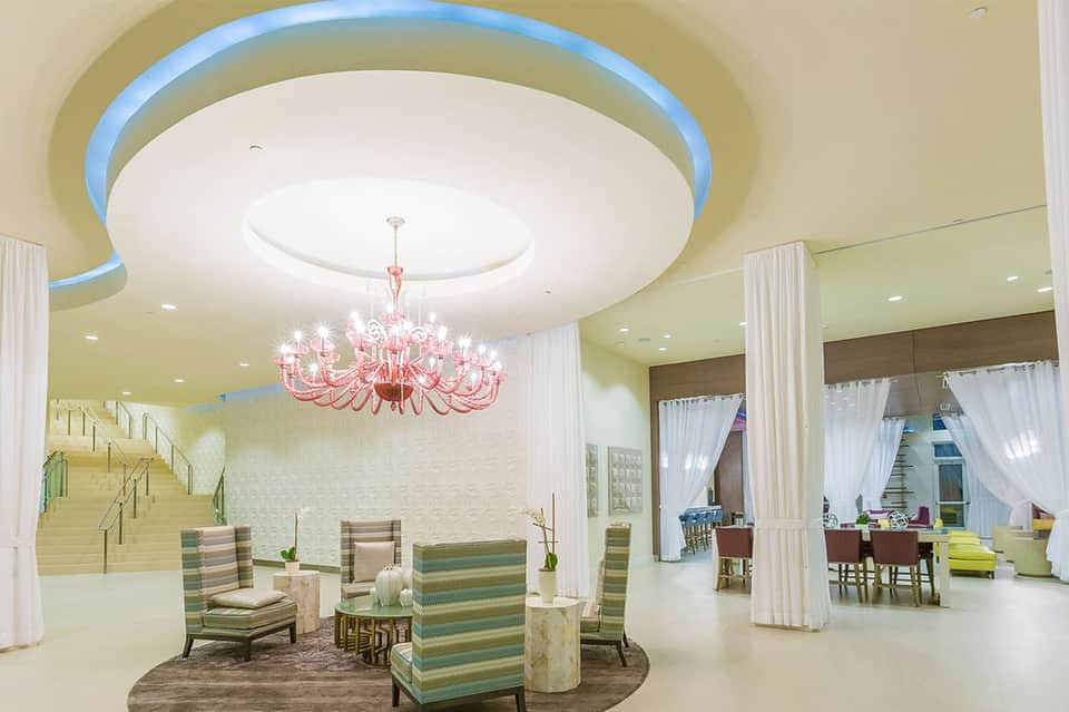 Modern lobby with yellow walls, pink chandelier, striped chairs, and curved ceiling with blue accent lighting