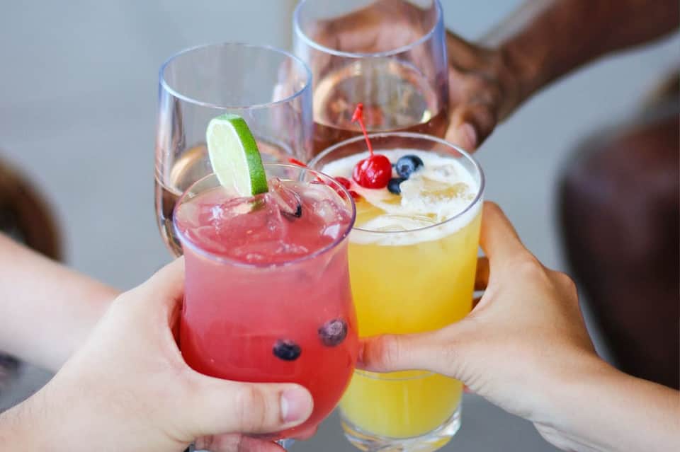 Hands holding colorful tropical cocktails with lime garnish and fruit toppings at a beach resort