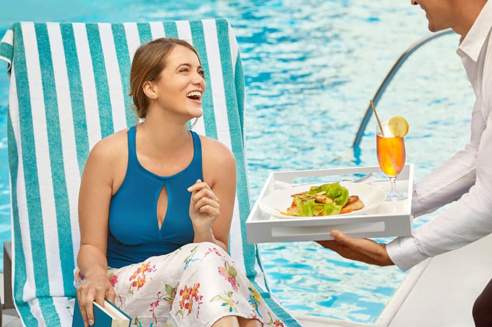 Woman relaxing on striped lounge chair by pool receiving beverage service