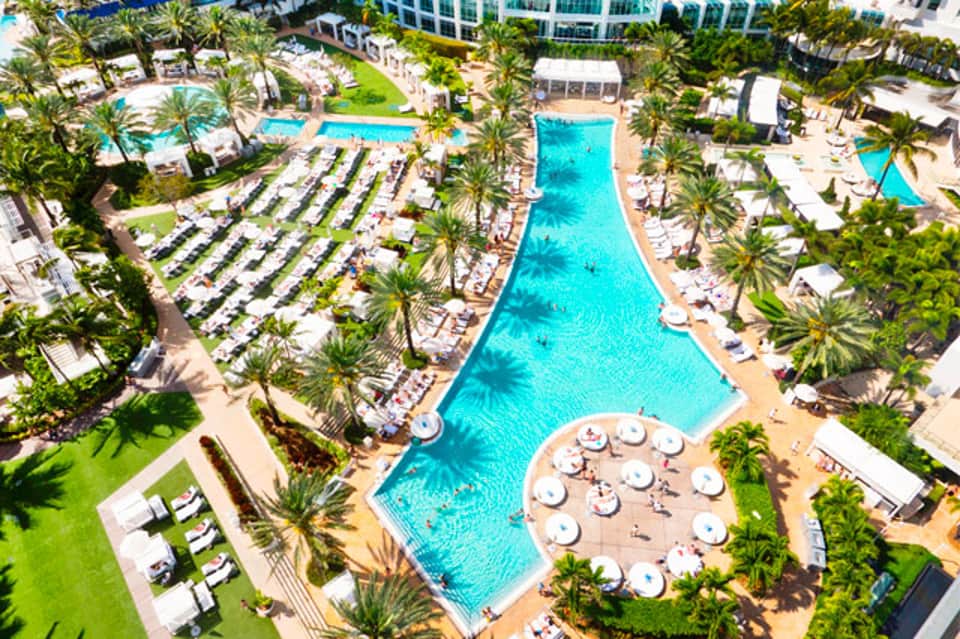 Aerial view of expansive tropical resort with multiple pools, lounge chairs, palm trees, and beachfront buildings
